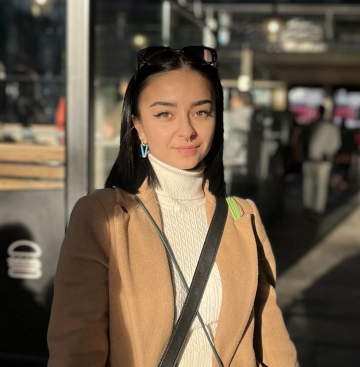 In the picture, you can see Martina Mellana standing on a street with a pleasant smile on her face, wearing a beige coat and a white turtleneck. 