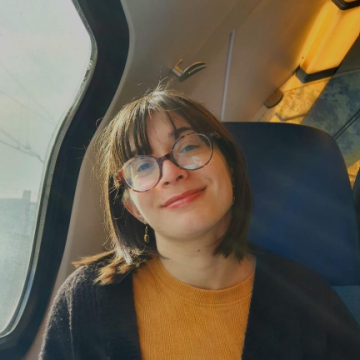 Samira Cruciani is smiling gently at the camera while seated in a train, wearing glasses, a mustard yellow top, and a dark cardigan. Sunlight filters through the window, creating a warm ambiance.